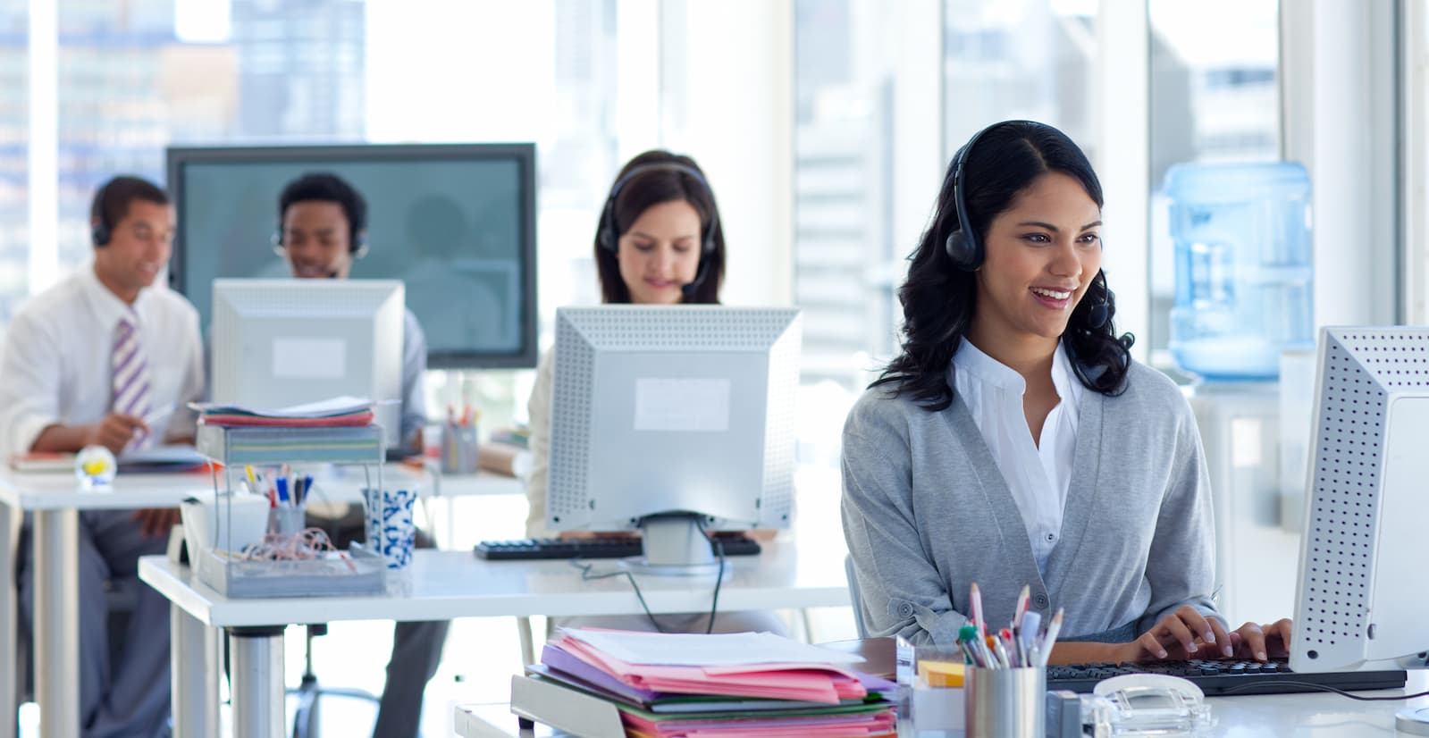 Customer service representatives working at desks in a bright office, with a focus on a smiling woman wearing a headset in the foreground.