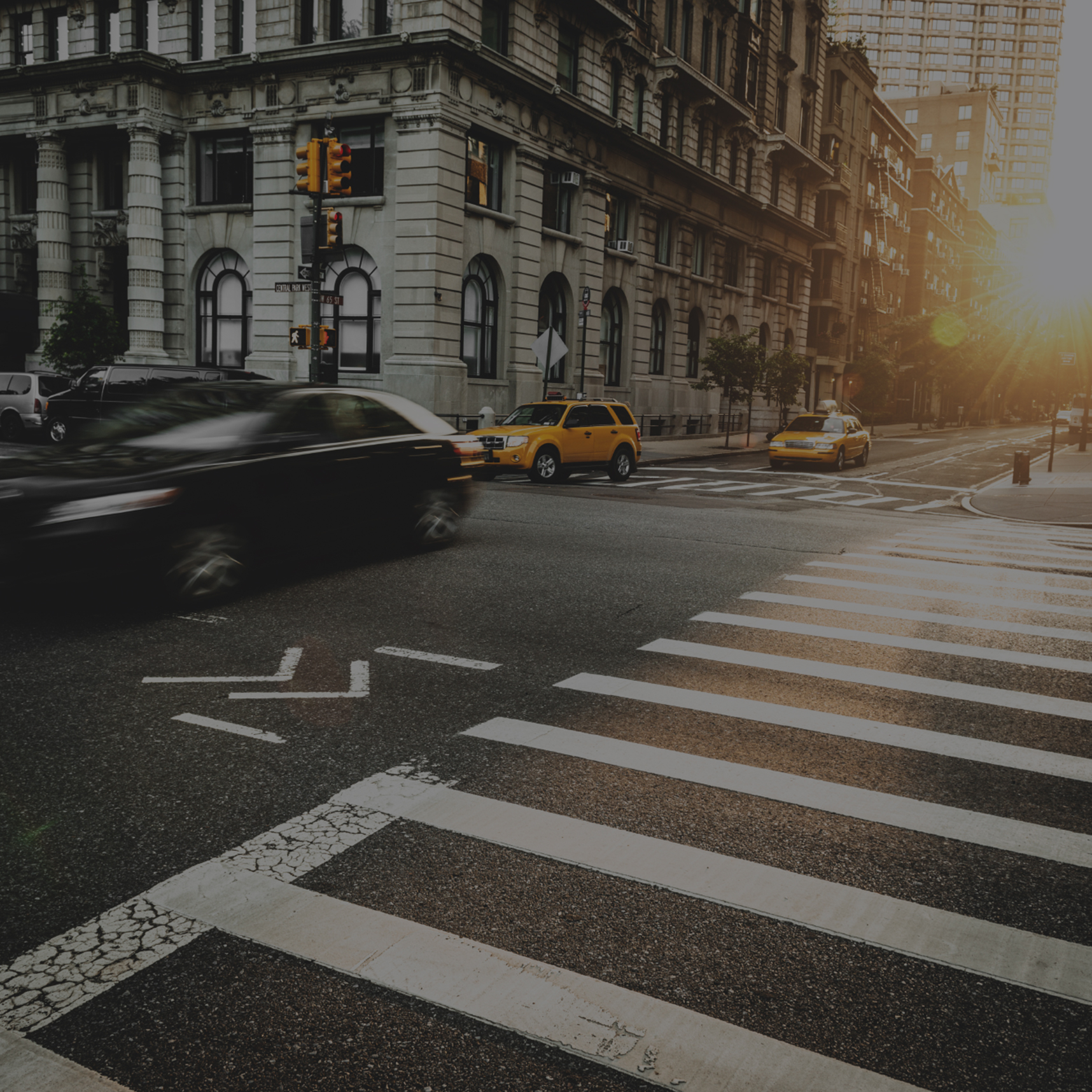 HeroforOtherIndustries-1716×1716 City street intersection at sunrise with blurred cars in motion, including a yellow taxi, and a pedestrian crosswalk in the foreground.