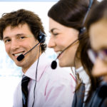Customer service representatives wearing headsets, smiling and working in a row at their desks.